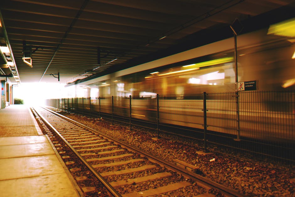 Inside a house or apartment near Southall Station, showing a section of a railway track with a paved platform on the left side, possibly part of a property entrance. The platform edge features concrete paving slabs, and there is a black metal fence separating the railway line from the building. In the background, a train is passing through at high speed, creating a motion blur effect with streaks of yellow and purple lights. Overhead, there is an underpass ceiling with visible beam structures and installed lighting, casting a subdued amber glow across the scene. To the left, a building with a store or commercial space can be partially seen, with its entrance or window area lit by internal lights. The scene captures the movement and urban environment typical of a busy rail station area, relevant to household removals, home relocation, and the logistics involved in furniture transport and packing during moves managed by companies like Man with Van Southall.