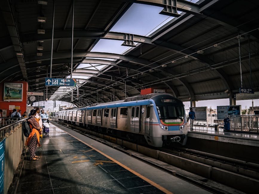 Inside a modern train station with a curved, partially transparent roof allowing natural light to fill the space. On the platform, several passengers, some carrying luggage or backpacks, are waiting for an arriving train. The train, painted in silver with blue, orange, and green stripe details, is currently stationary on the tracks, with its doors open. A few people are seen boarding the train, while others stand nearby. The station features safety railings along the platform edges, with signage overhead indicating directions to lifts and exits. The environment is clean and well-organized, with advertising screens and station information posters visible on the walls. This scene exemplifies the typical environment for home relocation or furniture transport involving train travel, coordinated by professional removals companies like Man with Van Southall, who manage logistical aspects of moving services efficiently.