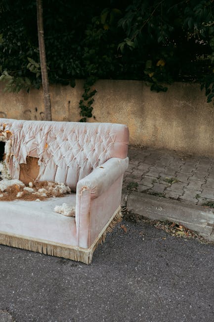 A partially damaged, vintage-style upholstered sofa with a light pink, velvet fabric adorned with button-tufted detailing on the backrest, is positioned outdoors on a paved driveway next to a stone curb. The sofa shows signs of wear and tear, with torn and missing fabric, revealing stuffing and underlying materials. It is situated near a concrete wall topped with greenery, including tree branches and leaves. In the background, a tall metal pole and a strip of pavement are visible, indicating the setting is outside a property, possibly awaiting disposal or removal. The scene is illuminated by natural daylight, and the sofa appears to be part of a furniture disposal or furniture transport process linked to home relocation or moving services, such as those provided by Man with Van Southall.