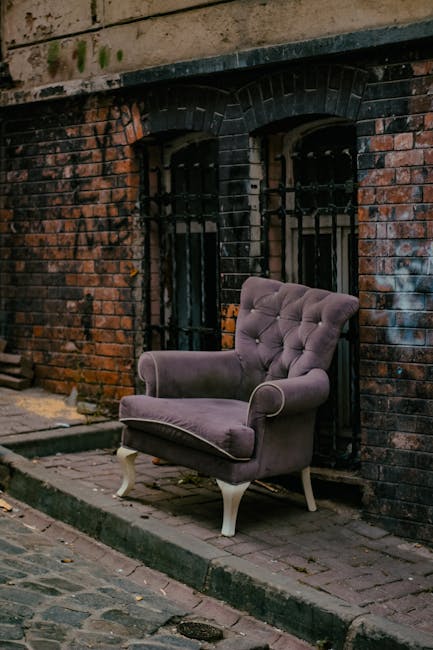 A purple velvet armchair with tufted upholstery and white wooden legs positioned on a brick pavement outside a building with a weathered brick wall. The chair is situated next to two small, barred windows with arched black metal bars set into the brickwork. The surrounding environment shows signs of aging with dark stains and patches on the brick wall. The scene appears to be part of a furniture removal process, with the chair possibly awaiting collection or disposal. This setting illustrates the typical urban environment where Man with Van Southall might assist with home relocation or bulky furniture disposal, involving loading and transport procedures within a moving services context.