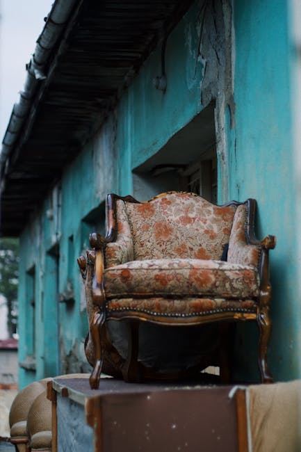 A partially damaged, vintage-style upholstered sofa with a light pink, velvet fabric adorned with button-tufted detailing on the backrest, is positioned outdoors on a paved driveway next to a stone curb. The sofa shows signs of wear and tear, with torn and missing fabric, revealing stuffing and underlying materials. It is situated near a concrete wall topped with greenery, including tree branches and leaves. In the background, a tall metal pole and a strip of pavement are visible, indicating the setting is outside a property, possibly awaiting disposal or removal. The scene is illuminated by natural daylight, and the sofa appears to be part of a furniture disposal or furniture transport process linked to home relocation or moving services, such as those provided by Man with Van Southall.