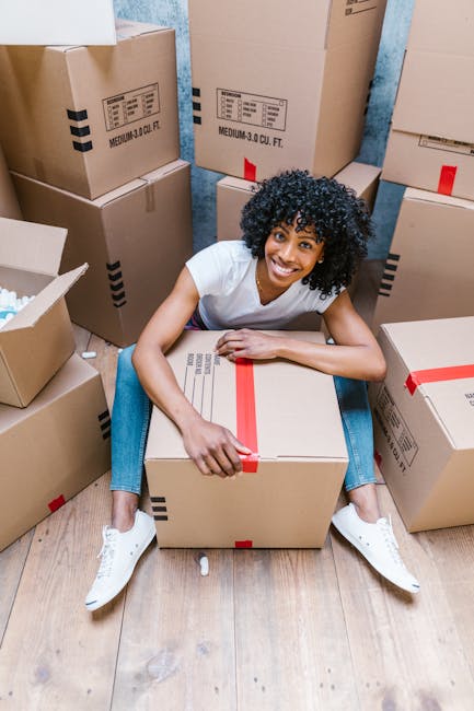 A woman with curly black hair, dressed in a white t-shirt and blue jeans, is sitting on a wooden floor surrounded by numerous cardboard boxes of various sizes, some sealed with red tape and others ope