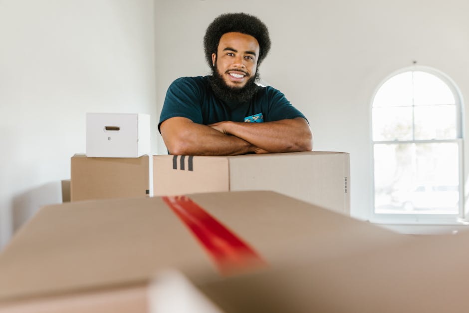 A woman with curly black hair, dressed in a white t-shirt and blue jeans, is sitting on a wooden floor surrounded by numerous cardboard boxes of various sizes, some sealed with red tape and others ope