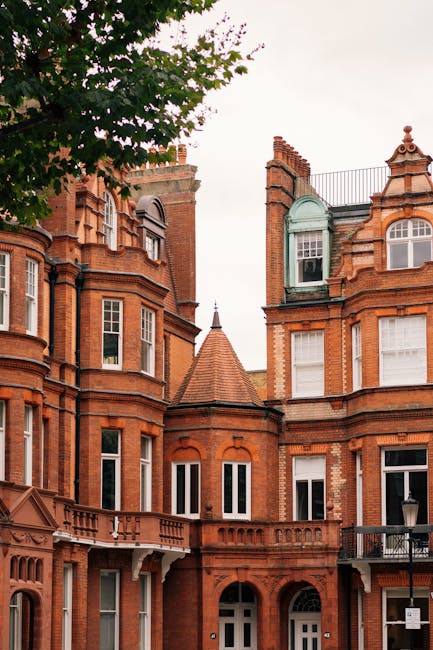 A row of historic red brick terraced houses with ornate architectural details, including bay windows, decorative stonework, and rounded turrets topped with conical roofs, situated on a street with a tree partially visible on the left side. The buildings feature large sash windows and balconies, consistent with traditional Victorian or Edwardian design. This exterior view, captured during daylight, illustrates classic residential architecture typical of the Southall area. Such properties often require careful packing and planning during home relocation or furniture transport, which companies like Man with Van Southall handle efficiently. The image highlights the external environment involved in moving services, emphasizing the importance of securing and protecting furniture and boxes during loading and transport processes, especially when navigating historic or multi-story buildings.