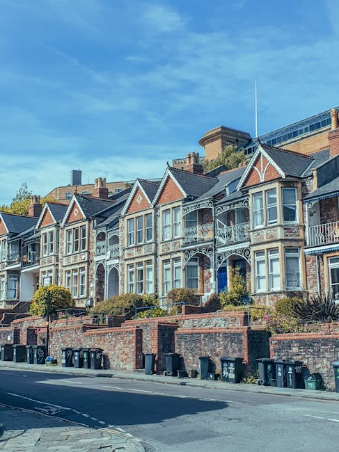 A row of Victorian-style terraced houses with intricate white decorative trim on the front facades, set above brick walls and steps leading to the entrances. The houses have large bay windows, some with small balconies, and pitched roofs with dark tiles. The street in front features a pavement with multiple black wheelie bins aligned along the curb. The scene is captured during daylight with a clear blue sky overhead. This residential area in Southall reflects typical urban architecture, which may require transportation permits for house removals or furniture transport services. [COMPANY_NAME], specializing in removals, often assists with the logistics involved in moving within areas like this, including the loading process and transport of household items and packing materials.