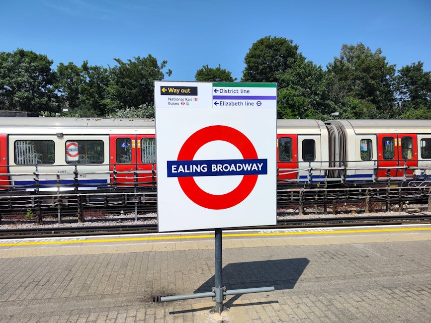 A railway platform at Ealing Broadway station with a large, upright sign displaying the station's name in white text on a blue background, positioned in front of a parked modern train with a silver body, red and blue accents, and several windows. The sign includes directional information for exit routes, bus services, and nearby districts such as Elizabeth line and National Rail, with a green and white color scheme. The platform surface is made of paving stones, and there are metal railings behind the train, along with green trees and a clear blue sky in the background. This scene showcases the typical environment for home relocation or furniture transport that may involve travel to or from Ealing Broadway, supported by professional removals services such as those provided by Man with Van Southall.