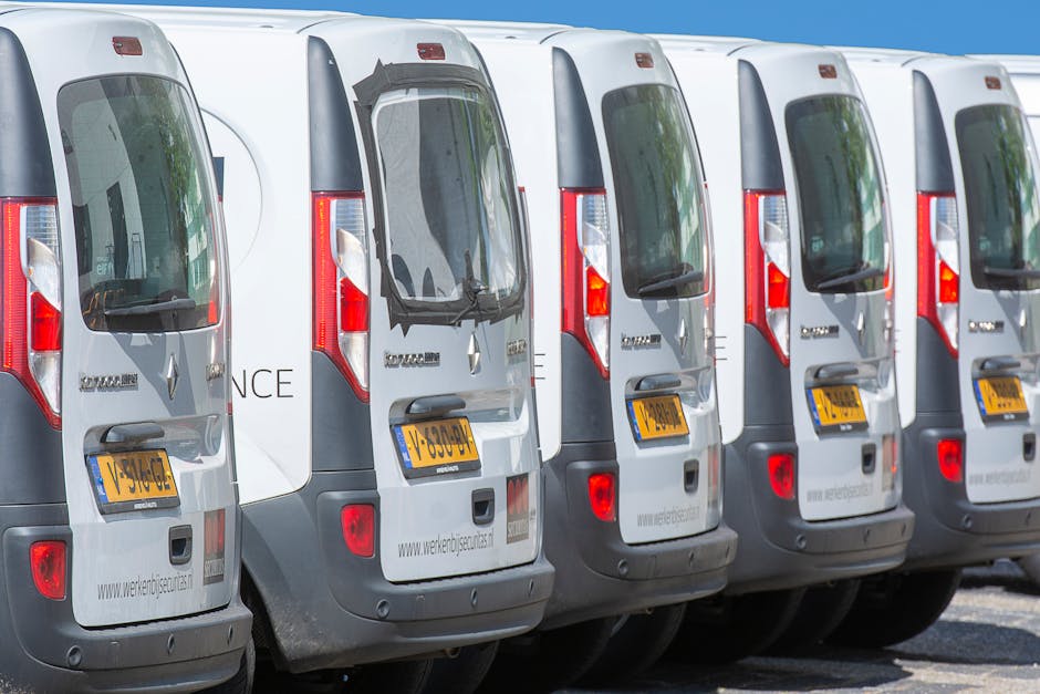 Six white and grey vans lined up side by side on a paved surface, with their rear doors facing the camera. The vehicles are part of a fleet used for furniture transport and home relocation services, as indicated by the website www.werkenjigseurla.com visible on the rear of each van. The vans are parked in an outdoor area under clear blue sky, with reflections of nearby buildings and trees visible in the rear window glass. Some vans display license plates with yellow backgrounds and black characters. No people or moving activities are visible in the image, which focuses on the parked vehicles, supporting the context of moving logistics and vehicle preparation managed by Man with Van Southall for house removals and professional relocation services.