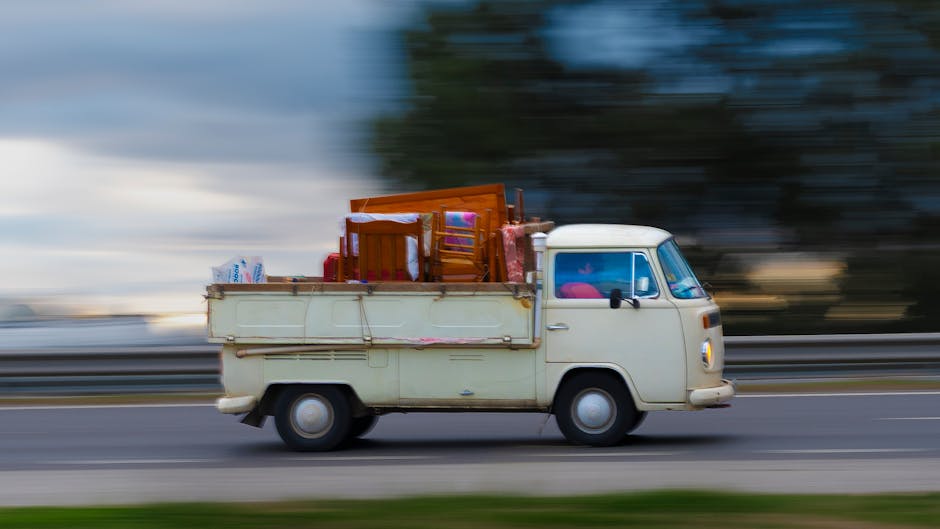 A vintage cream-colored van with a boxy design and rounded edges is moving along a multi-lane road during daytime with a slightly cloudy sky in the background. The van is loaded with various household furniture and packing materials, including a wooden chair, a small table, and cardboard boxes, which are secured in the open cargo area at the back. The furniture is wrapped in plastic or fabric for protection, and the boxes are stacked on top of the items. The vehicle appears to be in the process of transporting these belongings as part of a home relocation or furniture transport service. The background shows a blurred landscape of trees and sky, indicating motion, with the van approaching the right side of the image. This scene aligns with the logistics of house removals, where loading and securing household items are integral steps managed by Man with Van Southall, especially in the context of parking suspensions and van permits for Southall moves as described on the webpage.