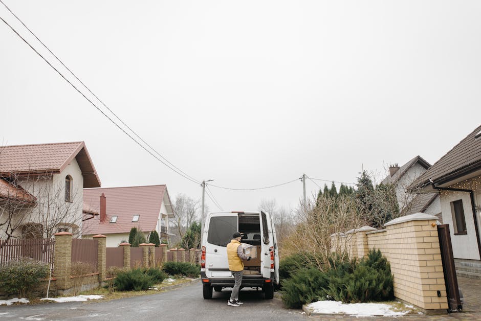 A man wearing a yellow jacket and a black cap is loading a cardboard box into the back of a white van parked on a residential street in Southall. The van is positioned near the doorstep of a house wit