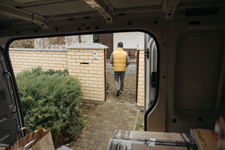 A man wearing a yellow jacket and a black cap is loading a cardboard box into the back of a white van parked on a residential street in Southall. The van is positioned near the doorstep of a house wit