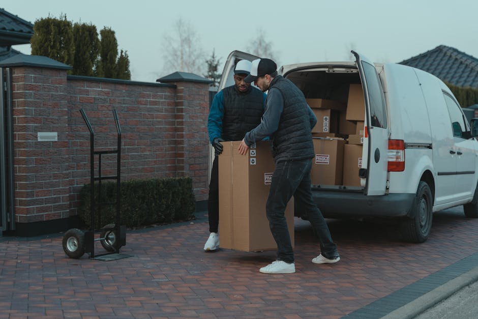 A man wearing a dark vest, blue long-sleeve shirt, black pants, white sneakers, and a blue cap is standing outside a residential property, leaning against the open door of a white van used for furnitu