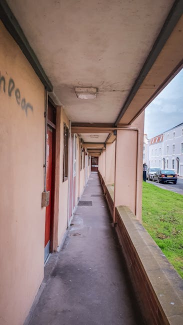 A long, narrow outdoor corridor on the upper floor of a residential building with a concrete ceiling and walls painted in a light tan colour. The corridor features a series of closed wooden doors, each with metal hinges and handles, and small, rectangular ventilation openings above some doors. The floor is made of concrete, and along the left side, there is a waist-high brick parapet with a wooden handrail for safety. To the right, the corridor opens to a small balcony area with a low brick wall, overlooking a street with parked cars and modern white residential buildings. Natural daylight illuminates the scene, and the corridor is clean but shows signs of wear, suitable for a home relocation or moving process where furniture and boxes might be transported through this outdoor passage. This setting corresponds with the services of Man with Van Southall, offering removals and relocation assistance within Southall and surrounding areas, especially when navigating narrow flat corridors.