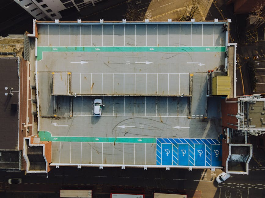 An aerial view of a rooftop parking area during daylight showing multiple designated parking spaces marked with white lines, including a row of standard parking bays, a section with blue disabled parking spaces, and a green-marked area for loading or bicycle storage. The parking lot surface is concrete with some rust stains and tire marks. A small white vehicle, possibly a van or car, is parked near the center, close to a covered loading zone with a corrugated metal roof supported by metal beams. Surrounding the rooftop, the tops of adjacent buildings are visible, with air conditioning units and other rooftop equipment seen along the edges. This image illustrates the logistics of home relocation and furniture transport planning, as it reflects the vehicle access points and parking arrangements typical for house removals in urban settings. The signage and markings support the understanding of parking access and loading zones used by [COMPANY_NAME] during packing and moving services, as highlighted on the Southall market removals page.