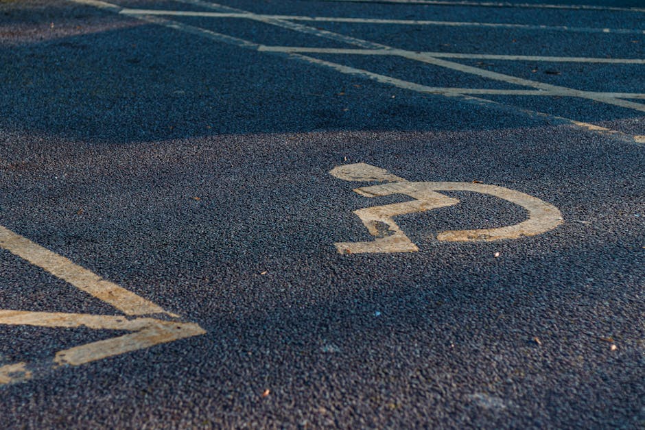 An aerial view of a rooftop parking area during daylight showing multiple designated parking spaces marked with white lines, including a row of standard parking bays, a section with blue disabled parking spaces, and a green-marked area for loading or bicycle storage. The parking lot surface is concrete with some rust stains and tire marks. A small white vehicle, possibly a van or car, is parked near the center, close to a covered loading zone with a corrugated metal roof supported by metal beams. Surrounding the rooftop, the tops of adjacent buildings are visible, with air conditioning units and other rooftop equipment seen along the edges. This image illustrates the logistics of home relocation and furniture transport planning, as it reflects the vehicle access points and parking arrangements typical for house removals in urban settings. The signage and markings support the understanding of parking access and loading zones used by [COMPANY_NAME] during packing and moving services, as highlighted on the Southall market removals page.