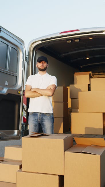 A young male mover with a beard, dressed in blue work overalls, is sitting at the open side door of a white van during a house removal process. He is holding and examining some paperwork, with a cardb