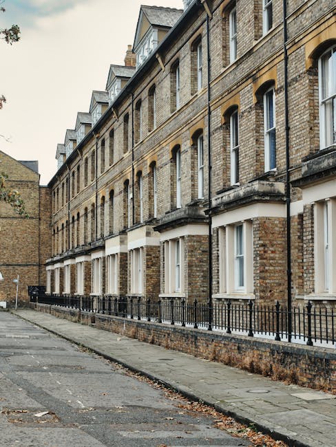 The image depicts a row of traditional terraced houses with brick facades and white-framed sash windows, situated along a pavement with a black metal fence running parallel to the buildings. The houses are multi-storey, featuring slate roofs with dormer windows at the top. Outside, there are several cardboard moving boxes stacked on the pavement, some wrapped in plastic for protection, indicating a home relocation process. Nearby, a wooden furniture dolly and moving blankets are visible, suggesting furniture transport preparations. The scene appears to be during daylight with overcast skies, and the area is quiet, with no people in visible motion. This setting aligns with the context of a house move, where [COMPANY_NAME] may be involved in the loading and transportation of household items, and the detailed environment reflects typical moving logistics in a residential neighbourhood on [PAGE_TITLE].