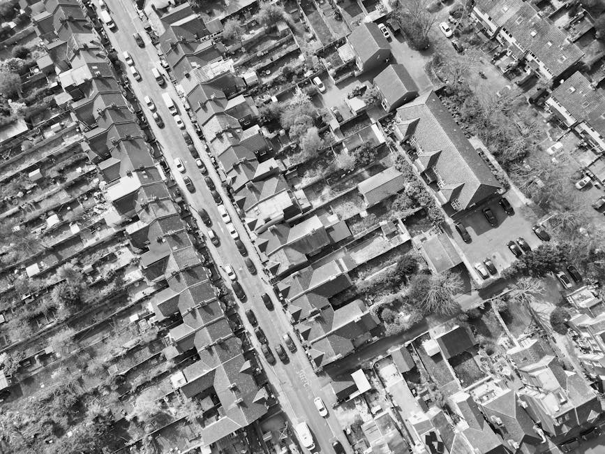 An aerial black-and-white photograph of a residential street showing closely packed terraced houses with pitched roofs, small front gardens, and narrow driveways. Several driveways are visible, some with parked cars, while others remain clear. The street runs parallel to the row of houses, with vehicles parked along one side. In the foreground, the side of a house with a small yard and trees can be seen, and a large van, likely belonging to Man with Van Southall, is parked on the pavement near an open driveway, suggesting a home relocation or furniture transport process in progress. The scene captures the organized arrangement of the homes and the surrounding environment, emphasizing the typical layout of a suburban area suitable for house removal services involving tight access and careful planning.
