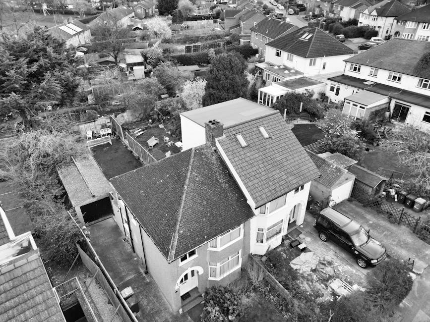 The image depicts a row of traditional terraced houses with brick facades and white-framed sash windows, situated along a pavement with a black metal fence running parallel to the buildings. The houses are multi-storey, featuring slate roofs with dormer windows at the top. Outside, there are several cardboard moving boxes stacked on the pavement, some wrapped in plastic for protection, indicating a home relocation process. Nearby, a wooden furniture dolly and moving blankets are visible, suggesting furniture transport preparations. The scene appears to be during daylight with overcast skies, and the area is quiet, with no people in visible motion. This setting aligns with the context of a house move, where [COMPANY_NAME] may be involved in the loading and transportation of household items, and the detailed environment reflects typical moving logistics in a residential neighbourhood on [PAGE_TITLE].