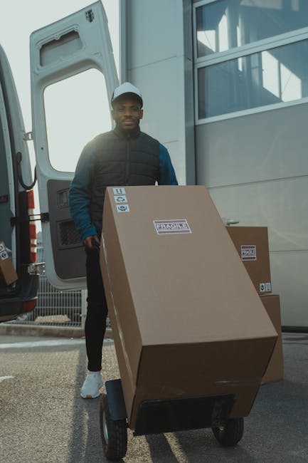 A man wearing casual clothing, including a black cap and padded jacket, stands beside a large cardboard box labeled 'Fragile' on a towable hand truck or cart. The box is positioned in front of an open vehicle door, with the interior of a van or truck visible behind him. Nearby, some additional cardboard boxes are stacked, also marked with 'Fragile' stickers. The scene appears to be outdoors, with part of a building or structure in the background and natural lighting illuminating the area. This image captures the process of home relocation, furniture transport, or packing and moving, consistent with services offered by Man with Van Southall, as they handle loading and moving household items in a professional manner.